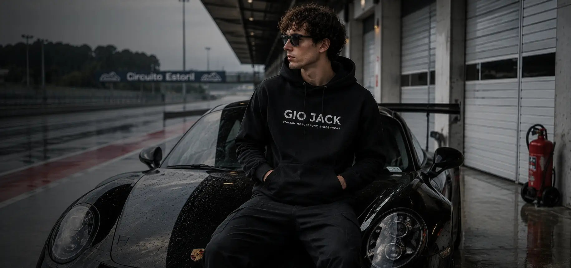 A man wearing a black hooded sweatshirt with 'GIO JACK' printed in white across the chest, sitting on the front of a black car in an outdoor urban setting.slider_item_najeNG